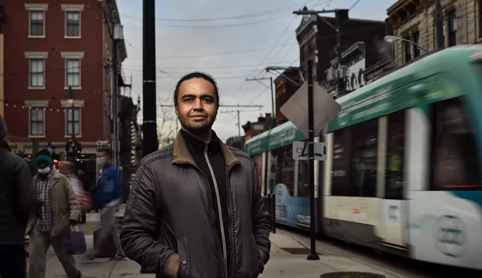 Stephen Gray stands on city sidewalk with street car passing by on right