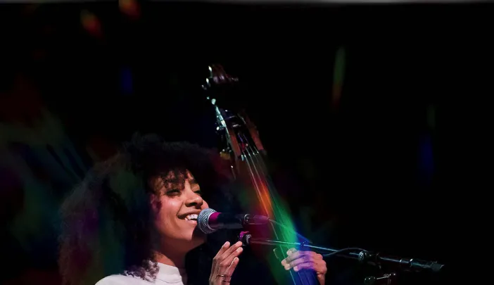 esperanza spalding, wearing a white jumpsuit with a "Life Force" logo, stands next to an upright bass