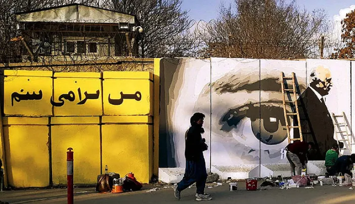 Along a city street, mural on a concrete wall shows two men shaking hands, with a large pair of watchful eyes in  the background.