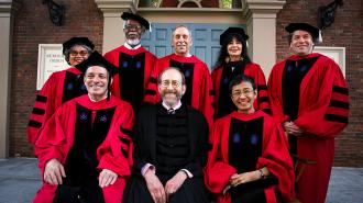 8 people in academic robes sit for a photograph in front on Memorial Church