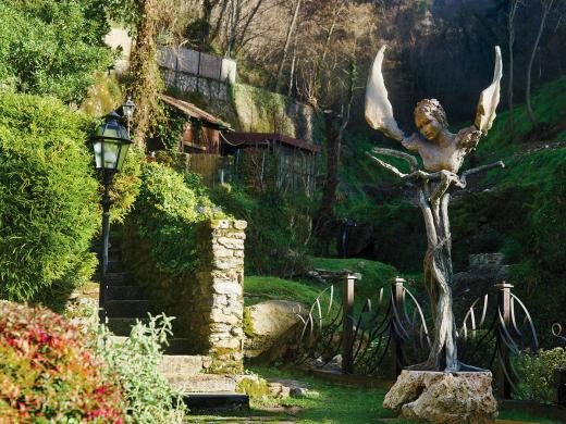 Bronze sculpture of a winged face and torso stands in a lush green garden