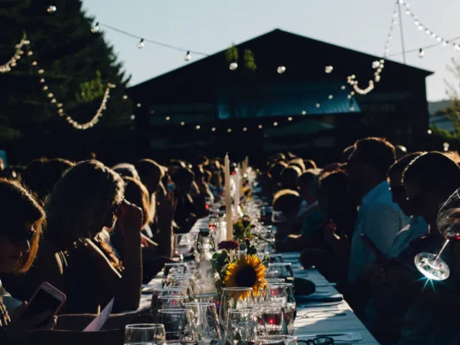 People gathered at a long, outdoor dining table set with wine glasses, candles, and flowers. 