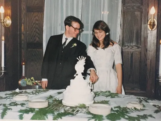 A bride and groom in formal attire cut a tiered cake at a candlelit wedding reception table.