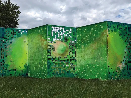 A six-foot-tall, five-panel folded screen stands in a field of grass next to the woods. It's painted different shades of green, with some squares cut out that represent digital pixels.