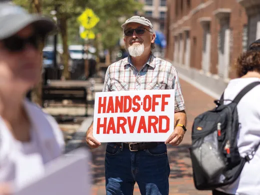 Man, standing in small group of people outside the courthouse, holding a sign that reads "HANDS OFF HARVARD" in red letters