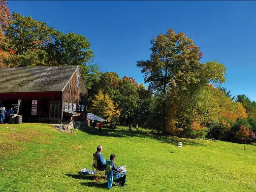 People sit in lawn chairs near a rustic barn at Cider Garden in New Salem on a sunny day.