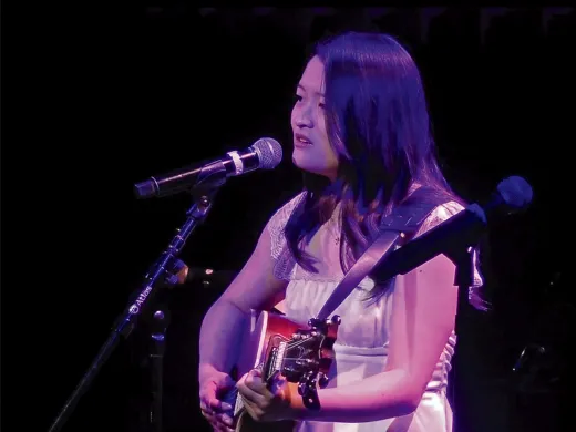 Julie Riew, wearing a white dress, playing guitar and singing into a microphone on stage.