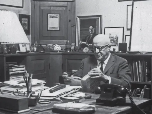 David McCord in suit reading a book at cluttered wooden desk in office filled with framed art and shelves.