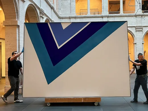 Two people moving large abstract painting with blue V-shaped design in museum courtyard.
