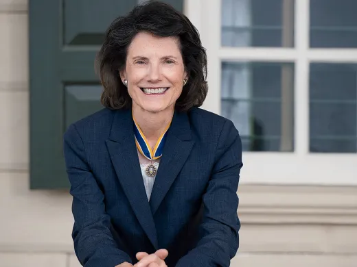 Anne Neal Petri in a navy suit leans on a wooden chair against an exterior wall of Mount Vernon..