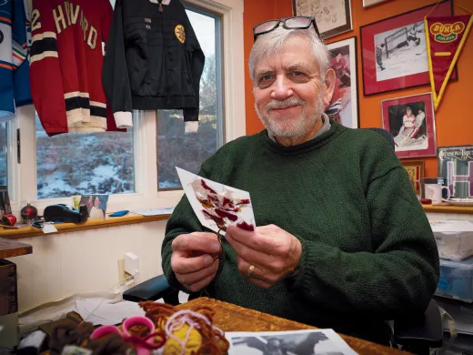 Older man in a green sweater holds a postcard in a warmly decorated office.