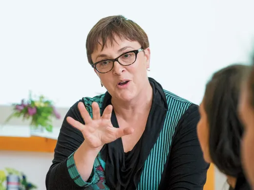 A woman in glasses gestures while speaking to two attentive listeners at a table.