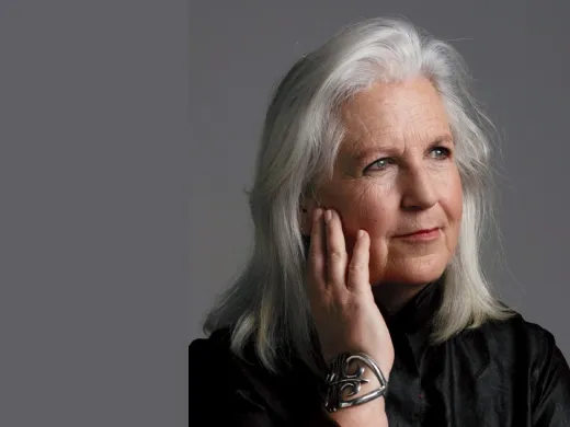 A woman with long, silver hair rests her chin on her hand, wearing a black top.