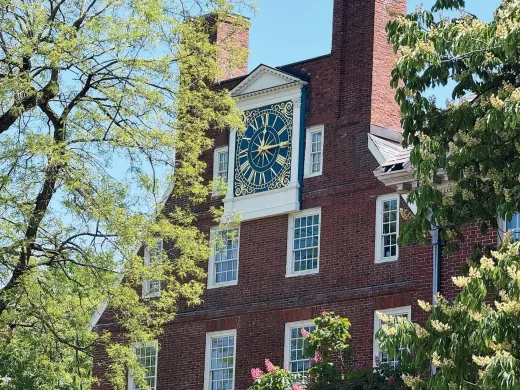 Massachusetts Hall at Harvard Red brick building with a large clock on top, surrounded by green trees.