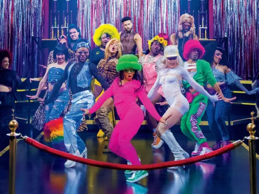 A vibrant group of dancers in colorful outfits poses on a stage with shiny decorations.