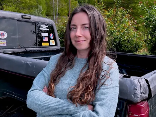 A woman with long hair stands confidently with crossed arms next to a pickup truck.