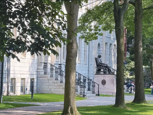 University Hall with steps and the John Harvard statue, surrounded by trees and a grassy lawn.