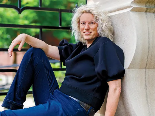 A woman with curly blonde hair sits casually against a stone wall, wearing a black top and jeans.