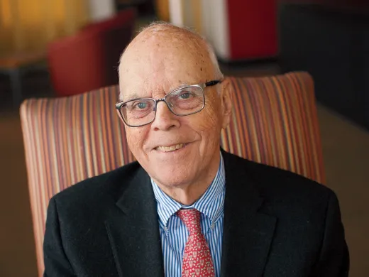 A smiling elderly man in glasses sits comfortably in a striped armchair.