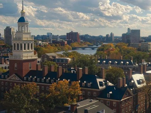Panoramic view of Harvard University with autumn foliage and a river under a cloudy sky.