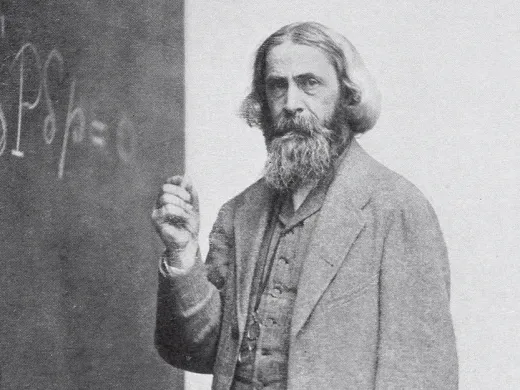 A man with a long beard and formal attire stands next to a chalkboard with an equation.