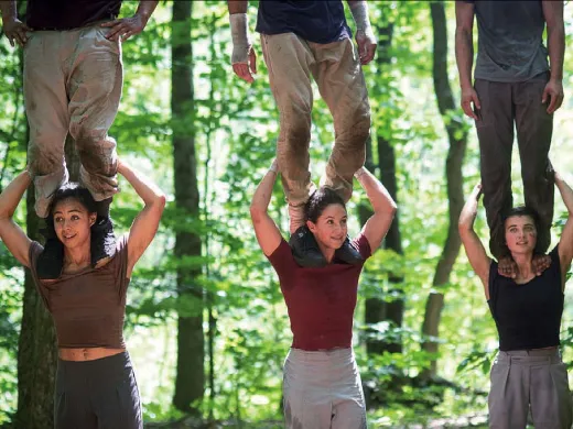 Four people perform a balancing act in a forest, with two standing on shoulders.