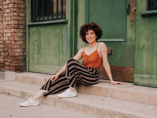Adriana Colón, a casually dressed, smiling young woman with curly brown hair, sits in a doorway.