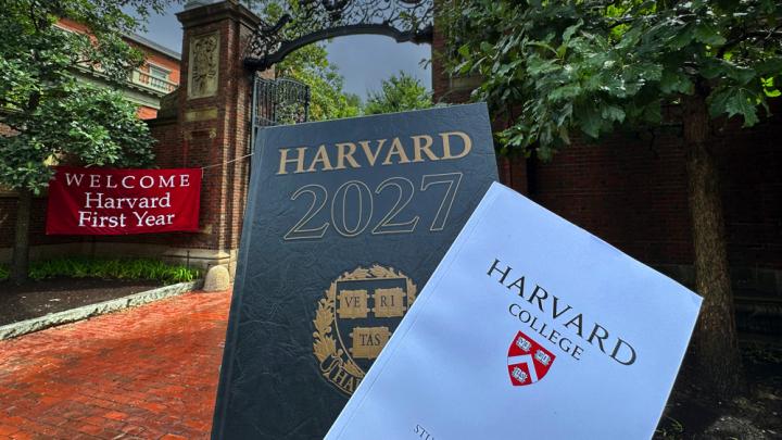 Harvard Gate in background with "Welcome Harvard First Year" red banner and in the foreground the Class of 2027 Class Book and Convocation program