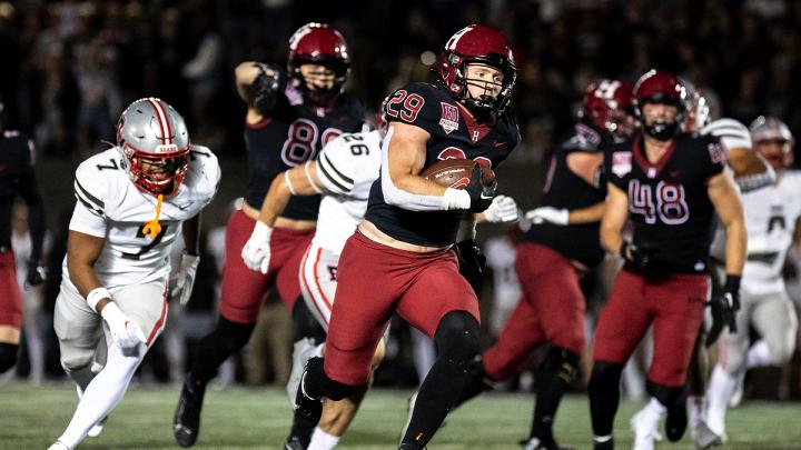 Harvard Football player 29 runs with football with Brown players running after along with other Harvard players