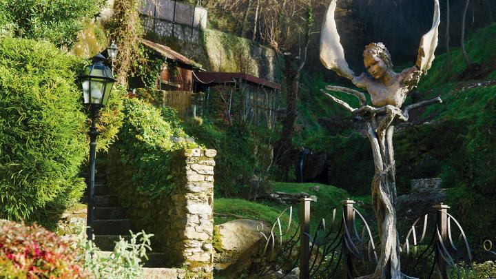 Bronze sculpture of a winged face and torso stands in a lush green garden