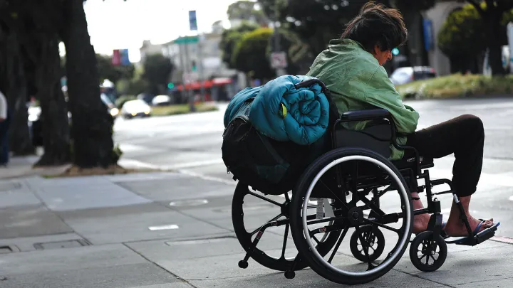 A man in a wheelchair with a rolled-up sleeping bag strapped to the back