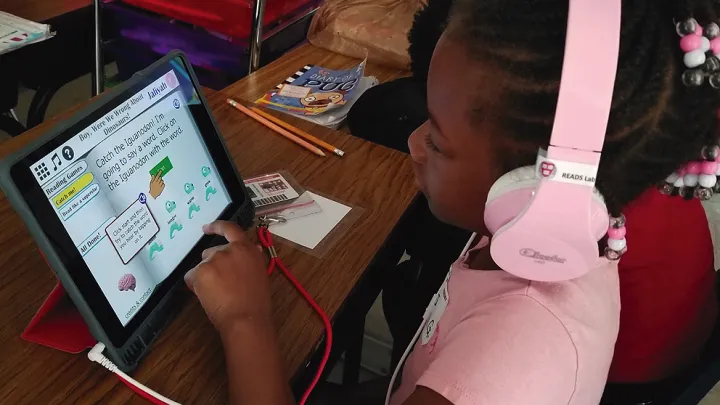 A young girl with pink headphones uses a tablet for an educational reading game at a classroom desk.