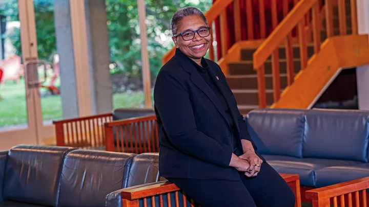 Smiling woman in glasses and a dark outfit seated indoors near a staircase and large windows.
