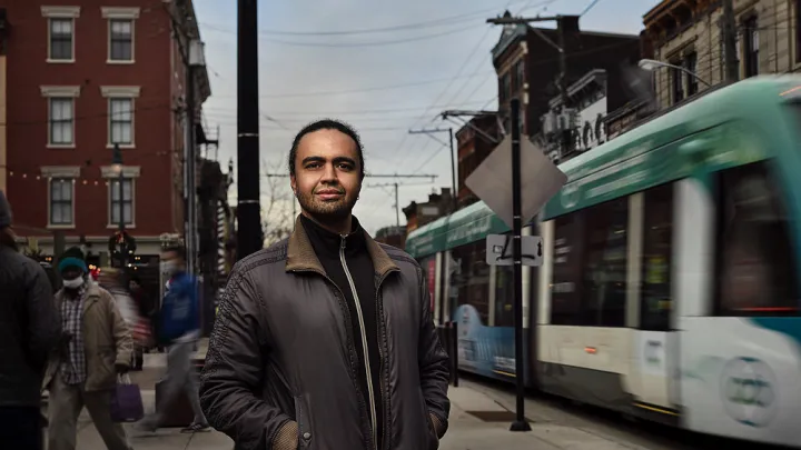 Stephen Gray stands on city sidewalk with street car passing by on right