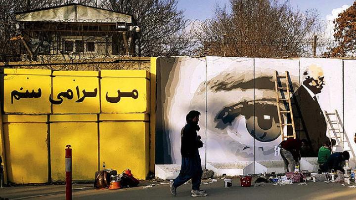 Along a city street, mural on a concrete wall shows two men shaking hands, with a large pair of watchful eyes in  the background.