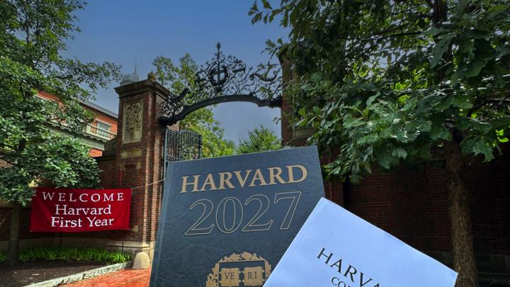 Harvard Gate in background with "Welcome Harvard First Year" red banner and in the foreground the Class of 2027 Class Book and Convocation program