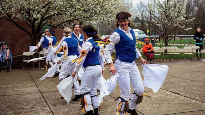 Morris dancers in white trousers and colorful vests wave handkerchiefs as they dance.