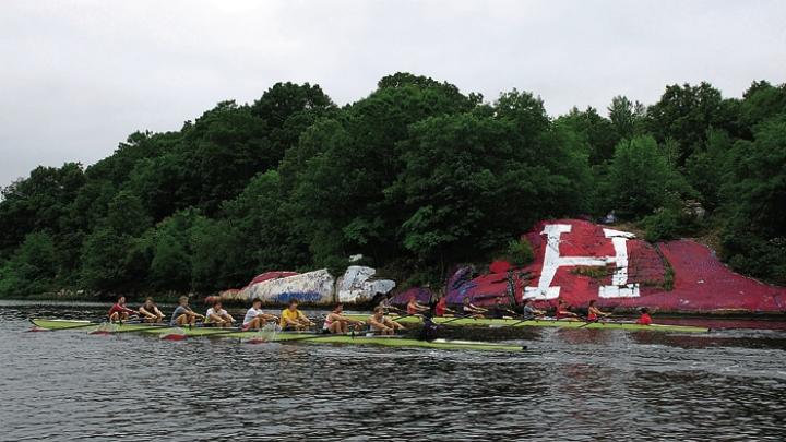 Two Harvard crews (right) row past “The rock,” long contested as a site for Harvard or Yale partisan artwork.