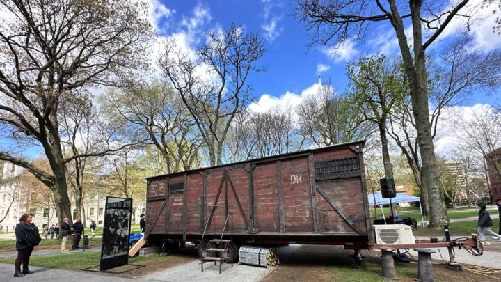A red-brown train car with boarded-up windows, harsh metal siding, and faded industrial German lettering is on display in Harvard Yard.