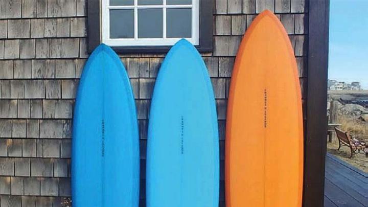 Two blue surfboards and an orange on rest on end against a weatherbeaten wall.