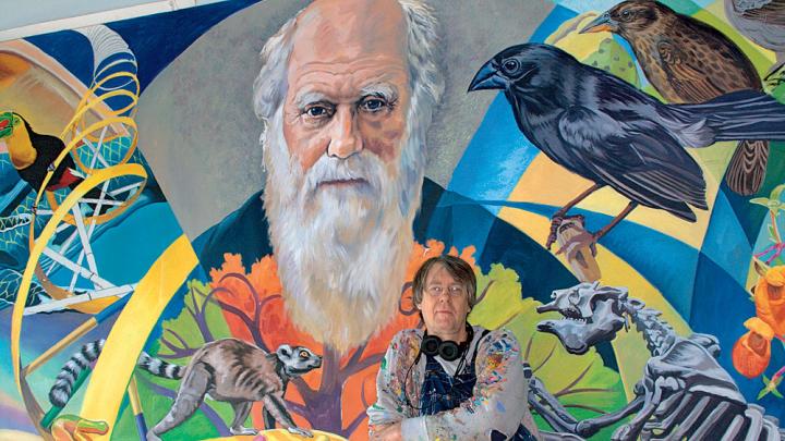 Fichter stands in front of <i>From Orchids to Octopi: The Evolution Mural,</i> which features Charles Darwin.