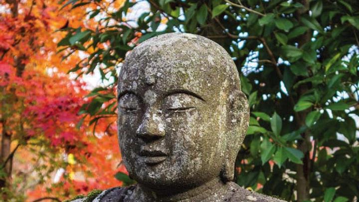 A stone statue of the Buddha appears against a background of colorful autumn leaves.
