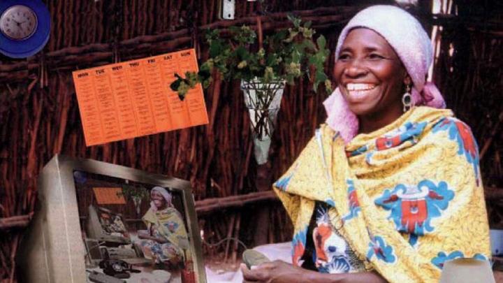 A grinning woman in traditional Nigerian dress sits cross-legged on the floor surrounded by modern devices, including a power strip, a land-line telephone, and a desktop computer displaying on its screen a duplicate image of the entire montage.