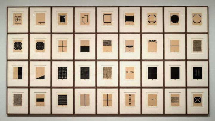 A grid of of 36 abstract black-and-white prints, each representing some aspect of the homes that artist Zarina has lived in, with Urdu and English inscriptions under each