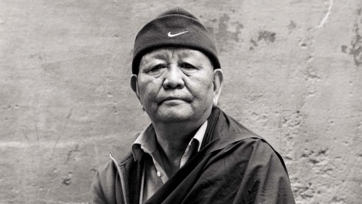 Black-and-white photograph of an older man with a robe draped over one shoulder and a zip-up sweatshirt poking out on the other, and a knit cap with a Nike logo, looks straight at the camera.