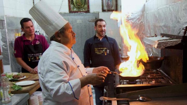 Two men look on while a chef cooks a flaming dish over a stove.