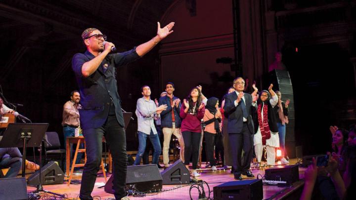Singer Ali Sethi gestures toward the audience at Sanders Theatre, while students dance on stage behind him.