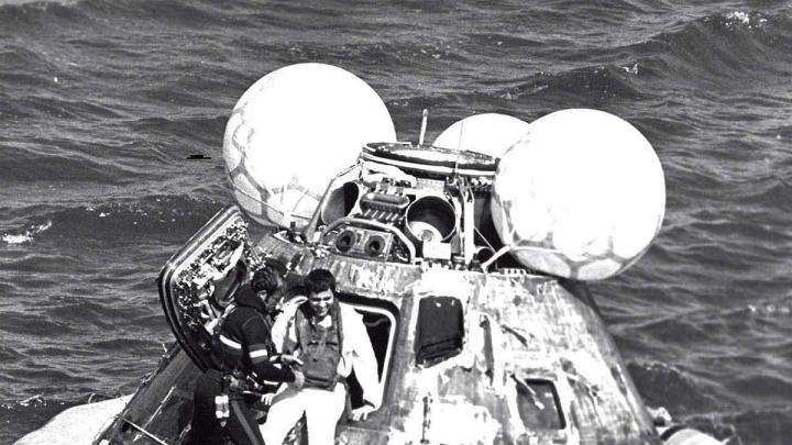 A man in a wetsuit helps an astronaut out of an Apollo capsule floating in the ocean