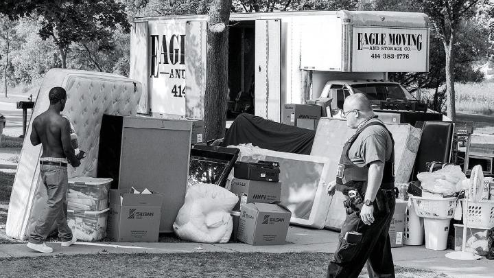 A deputy sheriff supervises as an evicted tenant carries belongings to the curb.