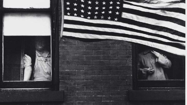 Robert Frank photograph of partially obscured people looking out of windows behind a large American flag